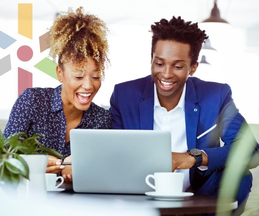 Two colleagues sitting at a table, smiling as they review something on a laptop in a modern office.
