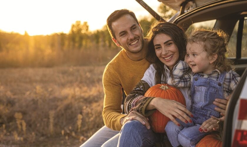 Parents and child sitting in the back of car trunk, surrounded by pumpkins.