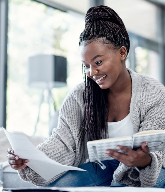 A young Black woman in a gray cardigan reviewing notes in a bright living room, symbolizing confident financial learning.