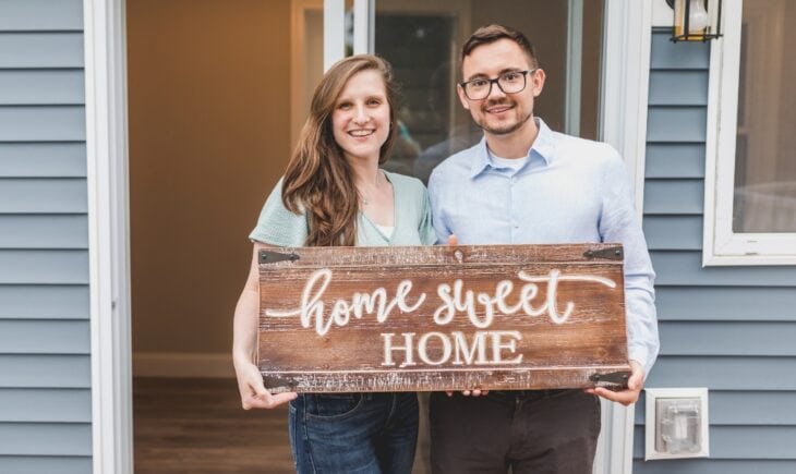 A smiling young couple stands in the doorway of a house with blue siding, holding a rustic wooden sign that reads "home sweet HOME" in white script and block lettering.