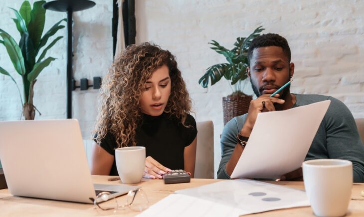 Two adults work through a household budget at a table with a laptop, calculator, and paperwork, practicing “know your numbers” budgeting basics for financial confidence.